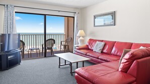 Een hotelkamer met een rode leren bank, een witte tafel, een televisie en een balkon met uitzicht op zee.
