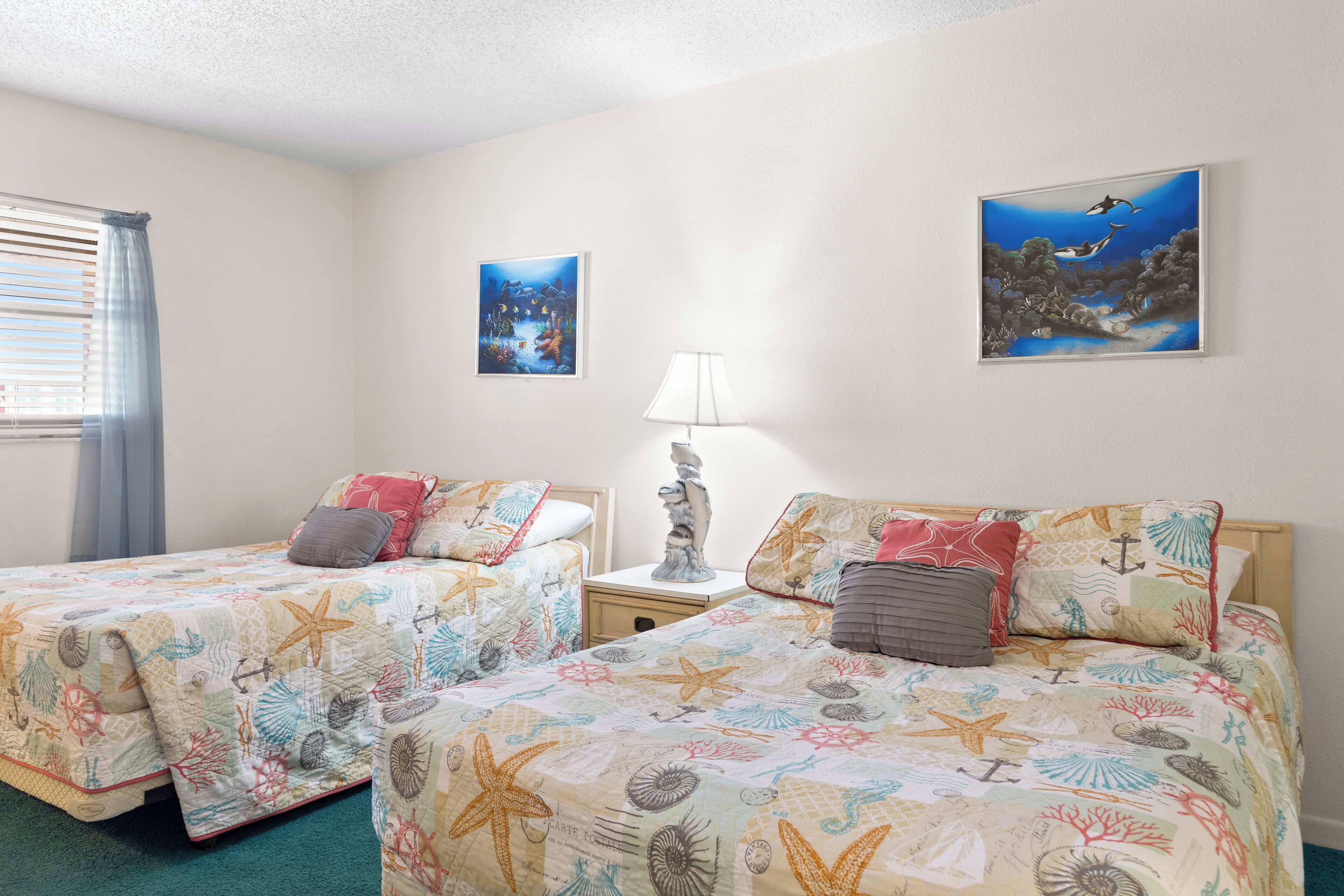 A room with two beds, each with a starfish-themed bedspread, a nightstand with a lamp, and two wall-mounted paintings depicting underwater scenes.