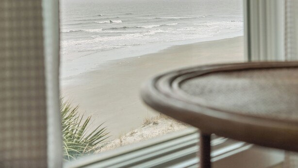 Vista de una playa a través de una ventana, con una mesa redonda parcialmente visible al frente.