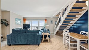 Un amplio salón con sofá, una mesa de centro y una mesa de comedor con sillas. Una escalera con una pared azul decorativa conduce a un nivel superior.