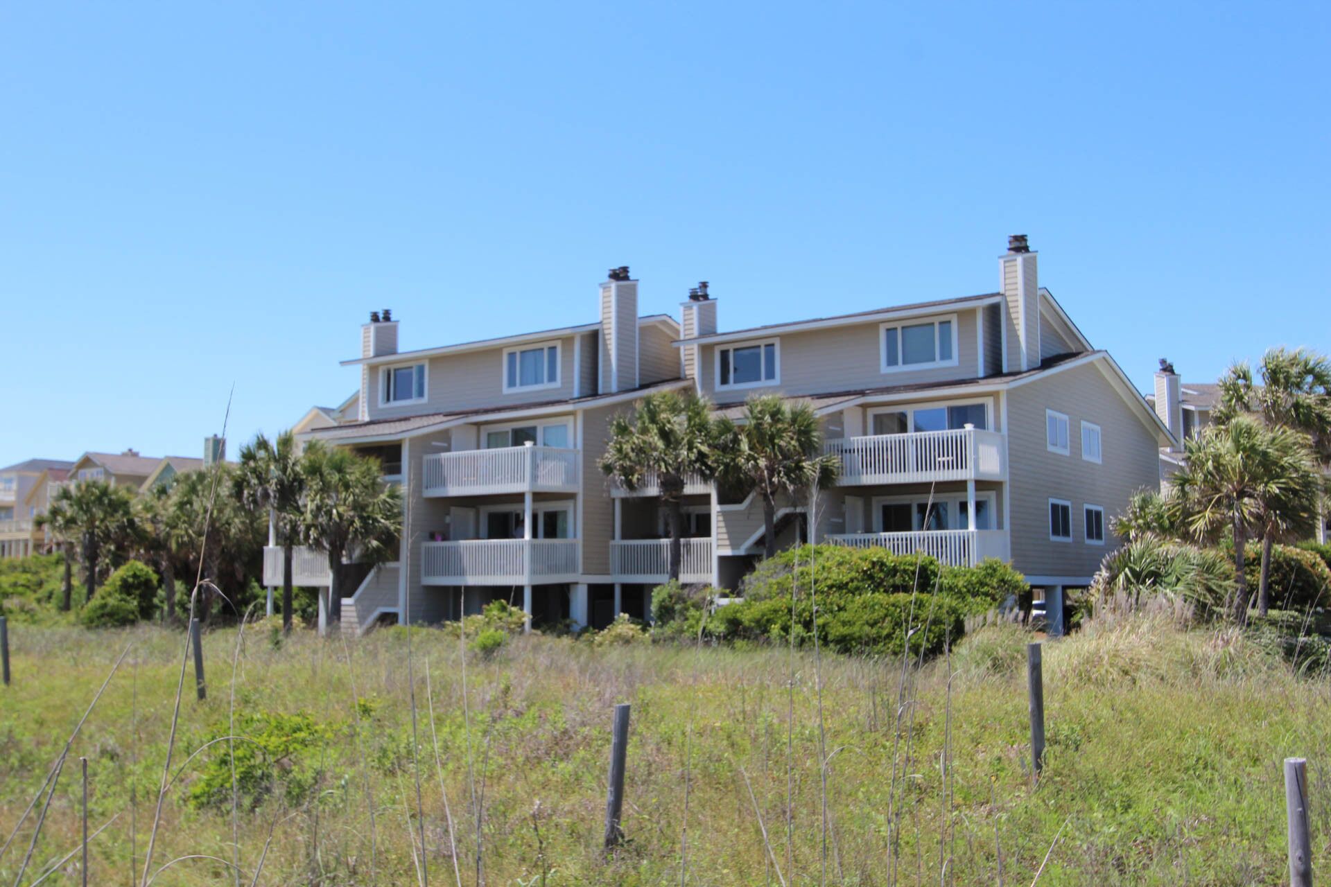A multi-story residential building with balconies, surrounded by vegetation and a clear sky.