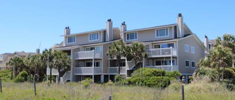 A multi-story residential building with balconies, surrounded by vegetation and a clear sky.