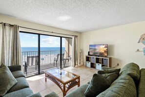 Un soggiorno con un'ampia porta scorrevole in vetro che dà su un balcone con vista sulla spiaggia e sull'oceano. Si vede una televisione su un mobile e un divano con cuscini.