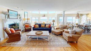 Een ruime woonkamer met een grote blauwe bank, beige fauteuils en een salontafel. Er is een open haard met een schoorsteenmantel en grote ramen met uitzicht op de oceaan.
