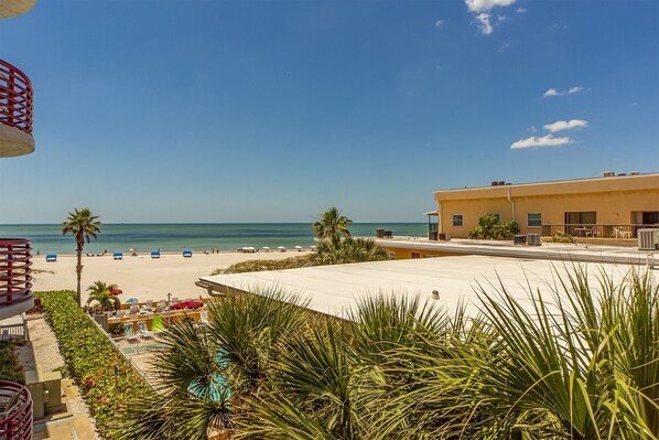 A beach view with a pool, palm trees, and a building with air conditioning units.