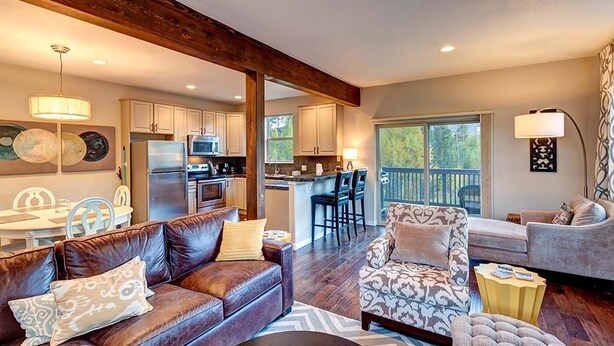 A spacious living area with a leather sofa, patterned armchair, and a dining table. A kitchen with modern appliances and a bar area is visible in the background. - Crossroads Snowshoe Townhome: Great Views, On Rec Path (Frisco)