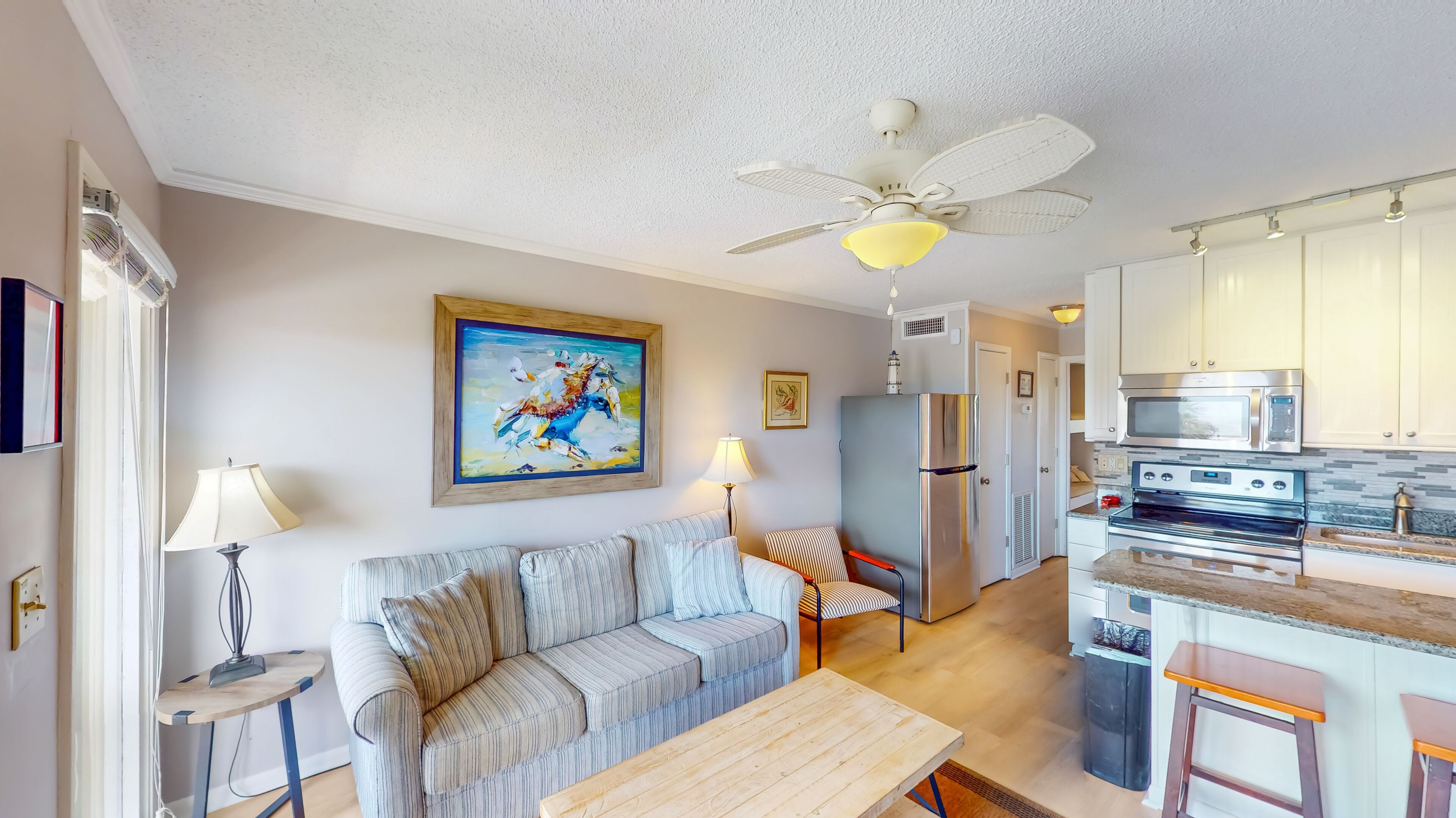 A living room with a sofa, a table, a ceiling fan, a painting, and a kitchen with appliances.