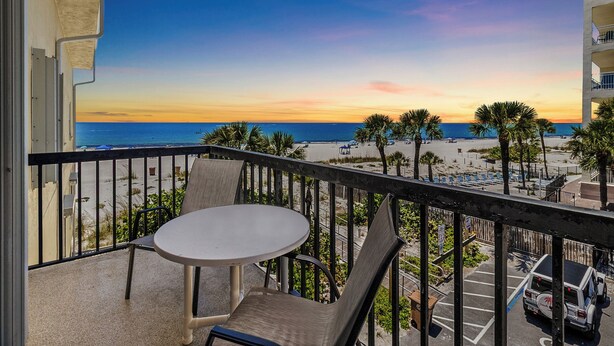 A balcony with a table and chairs, overlooking a beach with palm trees and a sunset.