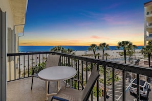 Een balkon met een tafel en stoelen, met uitzicht op een strand met palmbomen en een zonsondergang.
