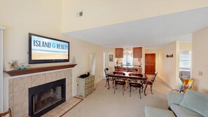 A living room with a fireplace, a dining area with a wooden table and chairs, a kitchen in the background, and a TV displaying 'ISLAND REALTY'. - Great home located near the beach with pool and tennis court access (Isle of Palms)
