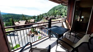 A balcony with a table and chairs, overlooking a residential area with houses and a forested mountain in the background.