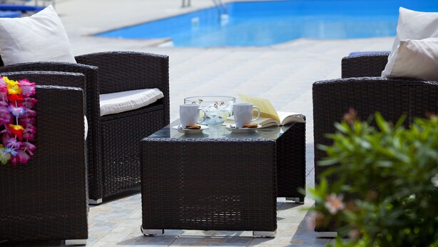 A poolside area with wicker furniture, a glass table with cups and a book, and a pool in the background.