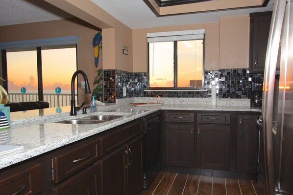 A modern kitchen with dark wood cabinets, a granite countertop, and a view of the sunset through the windows.