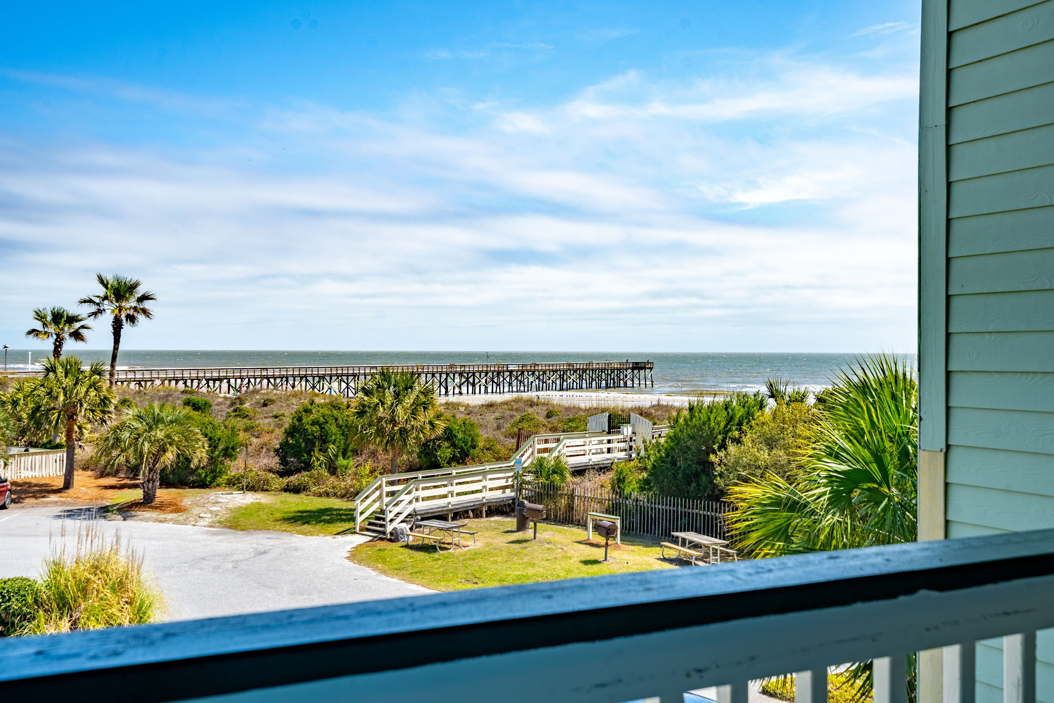 A view of a beach with a pier extending into the ocean, palm trees, and a fenced area with picnic tables.