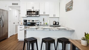 Een moderne keuken met witte kastjes, roestvrijstalen apparatuur en een marmeren aanrecht. Drie krukken en een houten tafel zijn aanwezig.