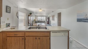 A kitchen with wooden cabinets, a dishwasher, and a dining area with a table and chairs. - Corner Unit Views John's Pass - Beach Place #204 (Madeira Beach)
