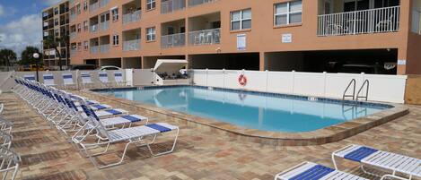 Una piscina con sillones de descanso y un edificio con balcones al fondo.