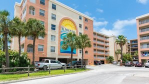 A multi-story hotel with a large sun mural on the facade, palm trees, and a parking lot with several vehicles. - Corner Unit Views John's Pass - Beach Place #204 (Madeira Beach)