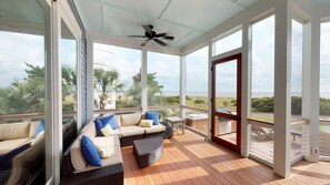 A covered outdoor seating area with wicker furniture and cushions, large windows offering a view of the ocean and greenery, and a ceiling fan. - Gorgeous home with sundeck overlooking the ocean and 18th hole on Wild Dunes (Isle of Palms)