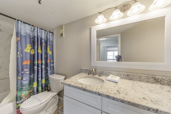 A bathroom with a toilet, a sink, a mirror, and a shower curtain with a fish pattern. - Beach View - Balcony - 3b 2b - Beach Place #305 (Madeira Beach)