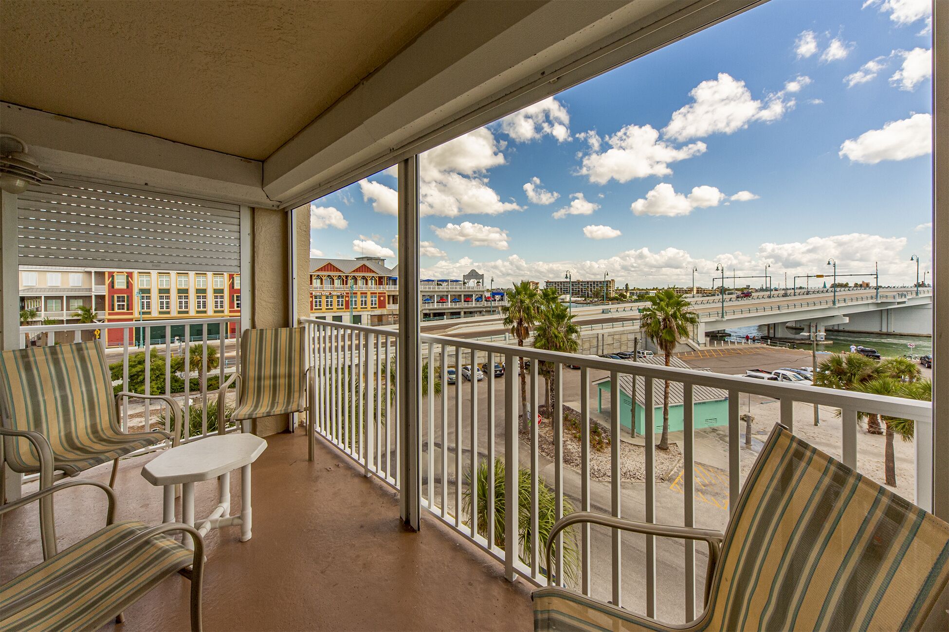 A balcony with striped chairs and a small table, overlooking a colorful building and a bridge.