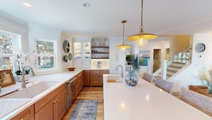 A modern kitchen with white countertops, wooden cabinets, and a central island. There are pendant lights, a built-in oven, and a view of the living area through an open door. - Beautifully Updated Wild Dunes Luxury House (Isle of Palms)