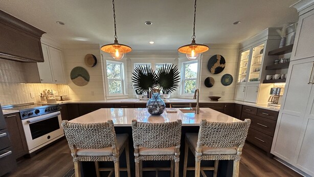 A kitchen with a central island, two hanging pendant lights, a built-in oven, and a dining area with three wicker chairs. - Beautifully Updated Wild Dunes Luxury House (Isle of Palms)