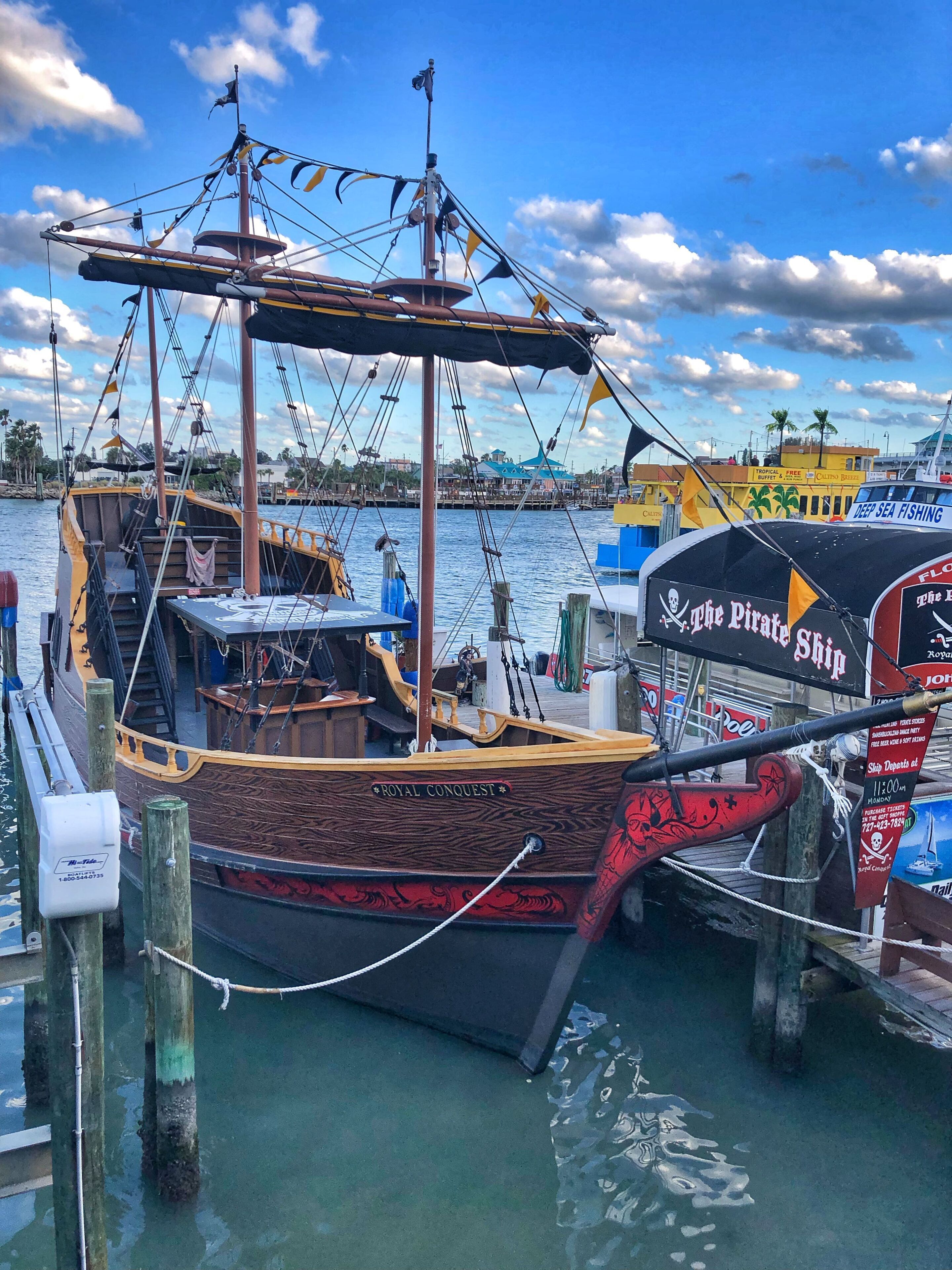A wooden boat named 'Royal Conquest' docked at a marina with a clear sky and a tropical backdrop.