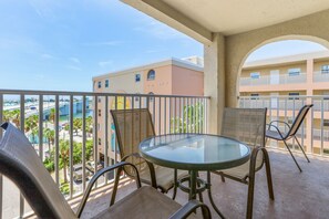 A balcony with a glass table and wicker chairs, overlooking a parking area and a building with a pink facade.