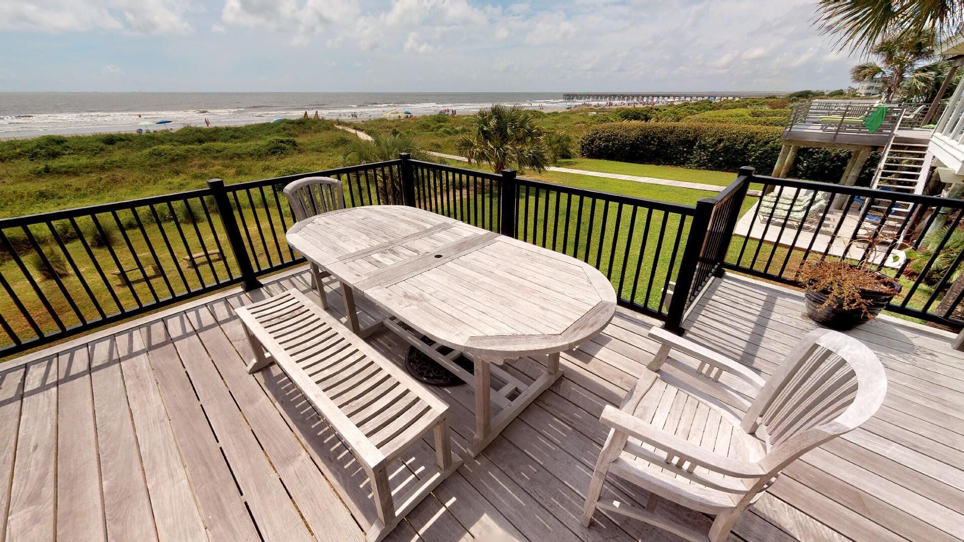 A wooden deck with a table and chairs, overlooking a beach and a pier.
