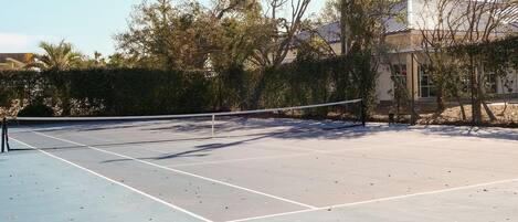 An outdoor tennis court with a net, surrounded by trees and a building in the background.