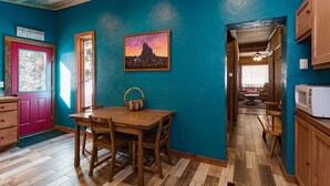 A cozy kitchen with a wooden table and chairs, a painting on the wall, and a view into the dining area.