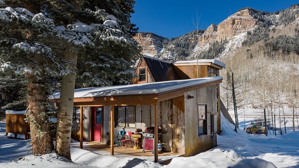 A wooden cabin with a glass door, surrounded by snow-covered trees and a mountainous backdrop.