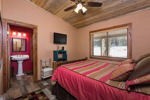 Un dormitorio con techo de madera, una cama con manta a rayas, un televisor en la pared y una ventana con persianas.