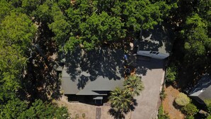 An aerial view of a property surrounded by dense greenery, featuring a large building with a sloped roof, a paved driveway, and a small parking area.