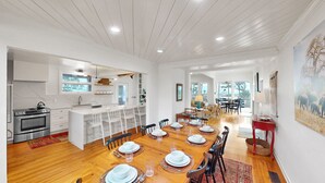 A spacious kitchen with a central island, a dining area with a wooden table set for six, and a view of the outdoors through large windows. - Updated Oceanfront home, large yard, private beach access, screened porch! (Isle of Palms)