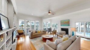 A spacious living room with a large sofa, a coffee table, and a fireplace. There are multiple windows providing natural light and a view of the outdoors.