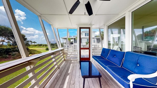 A covered porch with a blue sofa and a small table, overlooking a residential area with houses and greenery. - Updated Oceanfront home, large yard, private beach access, screened porch! (Isle of Palms)