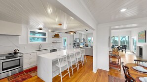 A modern kitchen with white cabinetry, a central island with bar stools, and a dining area with a fireplace. - Updated Oceanfront home, large yard, private beach access, screened porch! (Isle of Palms)