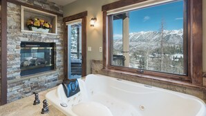 A bathroom with a stone fireplace, a large bathtub, and a window with a mountain view.