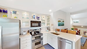 A modern kitchen with white cabinets, a built-in oven, microwave, and dishwasher. There is a dining area with wicker chairs and a table, and a painting on the wall. - Villa in Wild Dunes with golf and Lagoon views; close to pool (Isle of Palms)