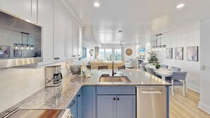 Cozinha moderna com bancadas de granito, eletrodomésticos de aço inoxidável e uma ilha central. Há uma área de jantar com mesa e cadeiras, e uma sala de estar com sofá e TV.