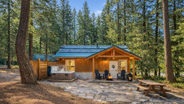 Une cabane dotée d’un patio en pierre, d’un bain à remous et d’un espace extérieur aménagé, le tout entouré d’arbres.