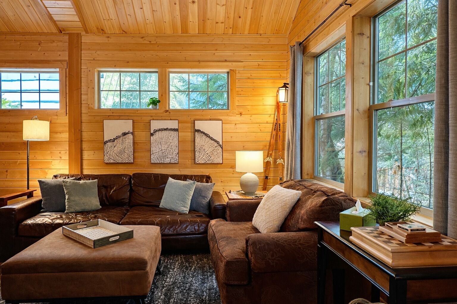 Een gezellig cabin interieur met een houten plafond, grote ramen, een leren bank en kunstwerken aan de wanden.