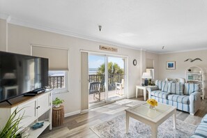 A living room with a TV, a sofa, a coffee table, and a balcony with a view of the beach.