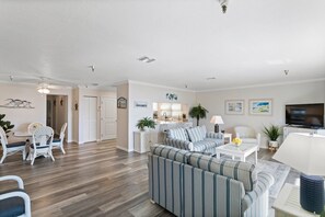 A spacious living room with a sofa set, dining area with chairs, and a kitchen in the background.