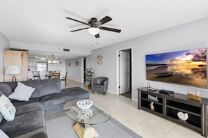 A modern living room with a grey sofa, a glass coffee table, a TV displaying a beach scene, and a ceiling fan.
