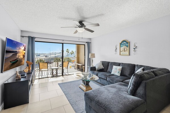 A modern living room with a grey sofa, a glass coffee table, a TV on the wall, and a balcony with a view of palm trees and a bridge. - Beachfront Complex - 2b 2b - Beach Place #203 (Madeira Beach)
