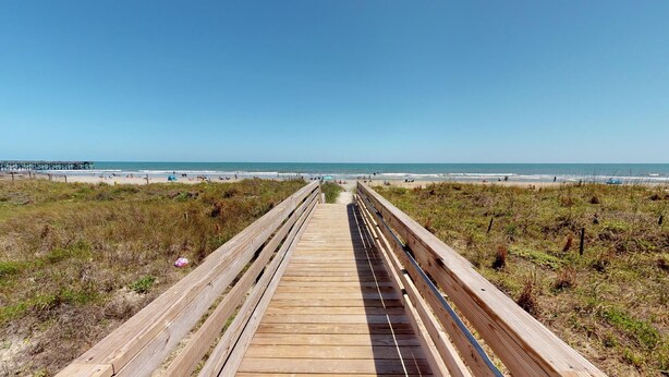 Una pasarela de madera que lleva a una playa, con un muelle al fondo.