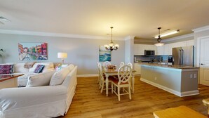 Una sala de estar moderna con un sofá blanco, una mesa de comedor con sillas y una cocina con armarios blancos y electrodomésticos de acero inoxidable.